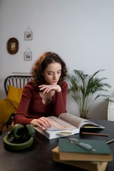 A young woman with curly hair reading a book indoors, surrounded by headphones and a cozy setting.