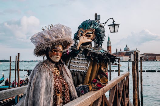 Two people in ornate Venetian costumes standing by water with iconic architecture in background.