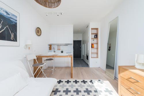 White couch placed on rug near table and drawer in stylish room with mirror and decorated shelves and long corridor with door