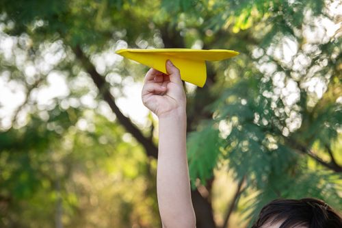 Free A child playfully raises a yellow paper plane against a lush green background. Stock Photo