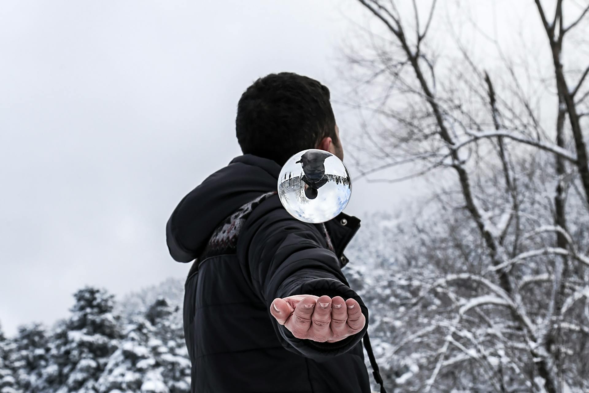 A man in winter clothing skillfully balances a glass orb reflecting the snowy forest surroundings.