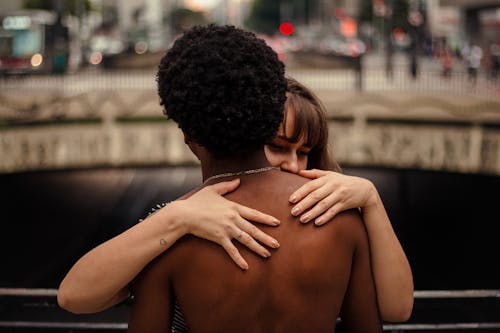Happy young gentle female embracing African American male warmly on blurred background of bridge