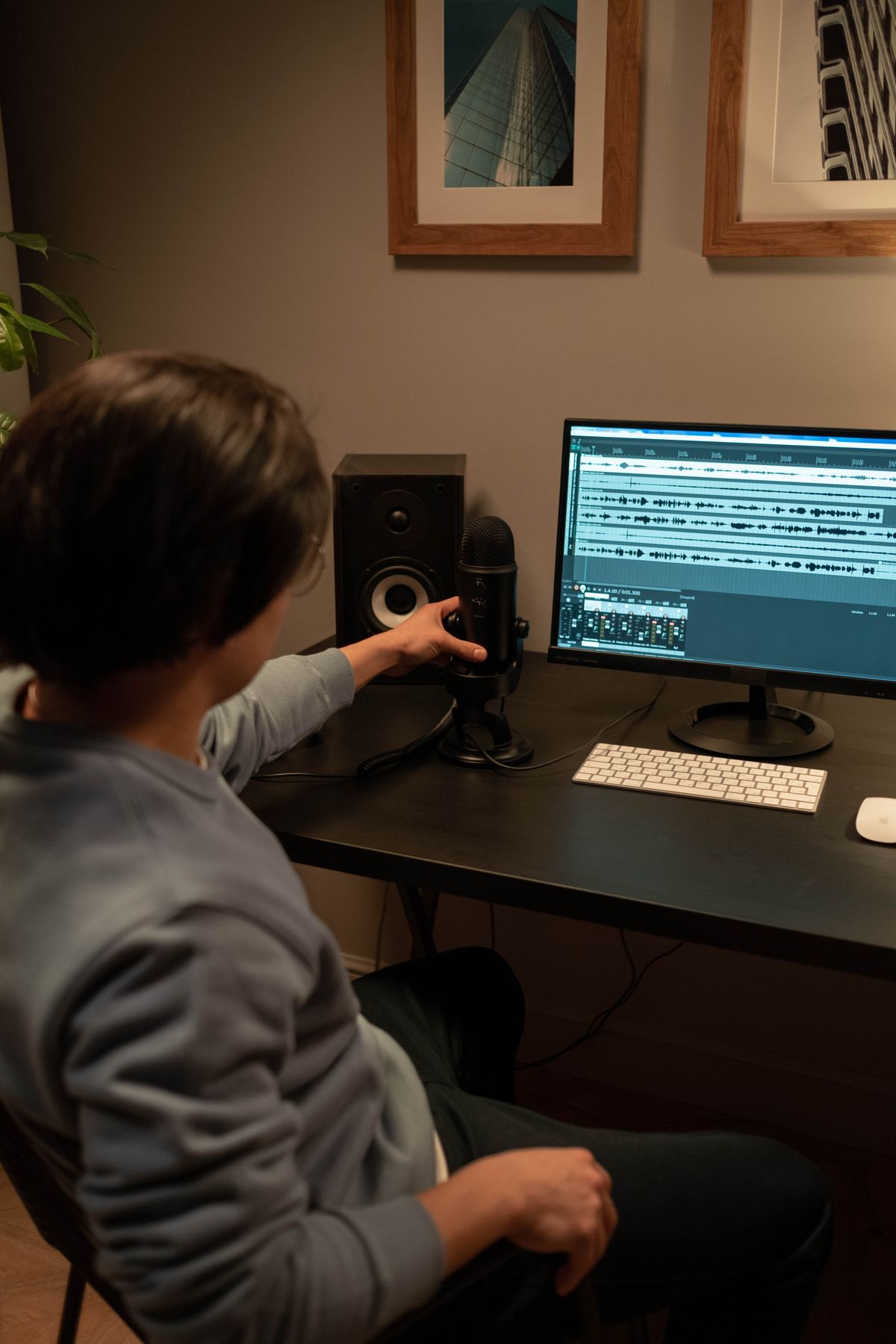 Person at a home desk adjusting a USB microphone next to a monitor showing audio waveforms