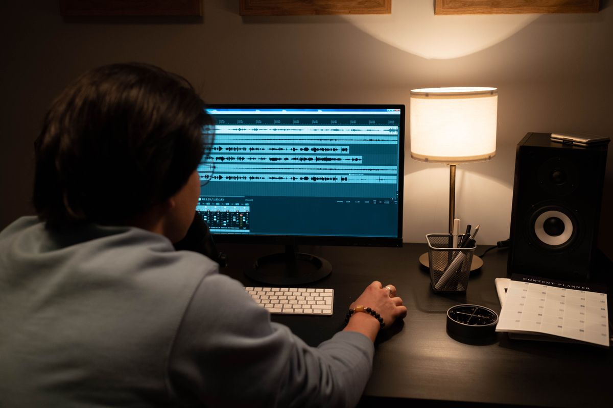 Persona de espaldas ante un monitor que muestra formas de onda de audio, con una lámpara cálida de escritorio y un altavoz de estudio