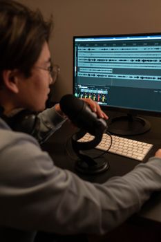 An adult man working in a recording studio, using a microphone and editing audio on a computer.