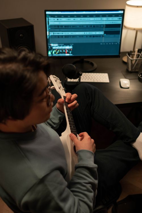 Free Adult playing ukulele in a cozy home studio, surrounded by modern recording equipment. Stock Photo