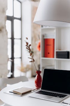 Stylish minimalist home office setup with a laptop, red vase, and decor on a white table.
