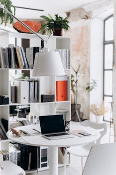Well-lit modern office with a laptop on a round table, surrounded by shelves and plants.