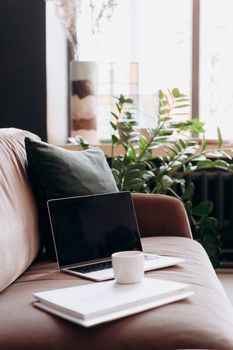 A cozy home office setup featuring a laptop, ceramic mug, and green plant on a couch by the window.