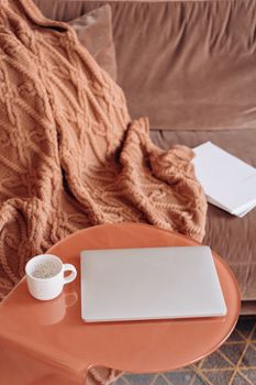 A warm and cozy home setting with a laptop and coffee on a wooden table next to a knitted blanket.