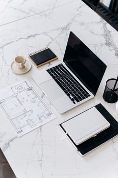 Overhead view of a modern desk with laptop, blueprint, and stationery on a marble surface.
