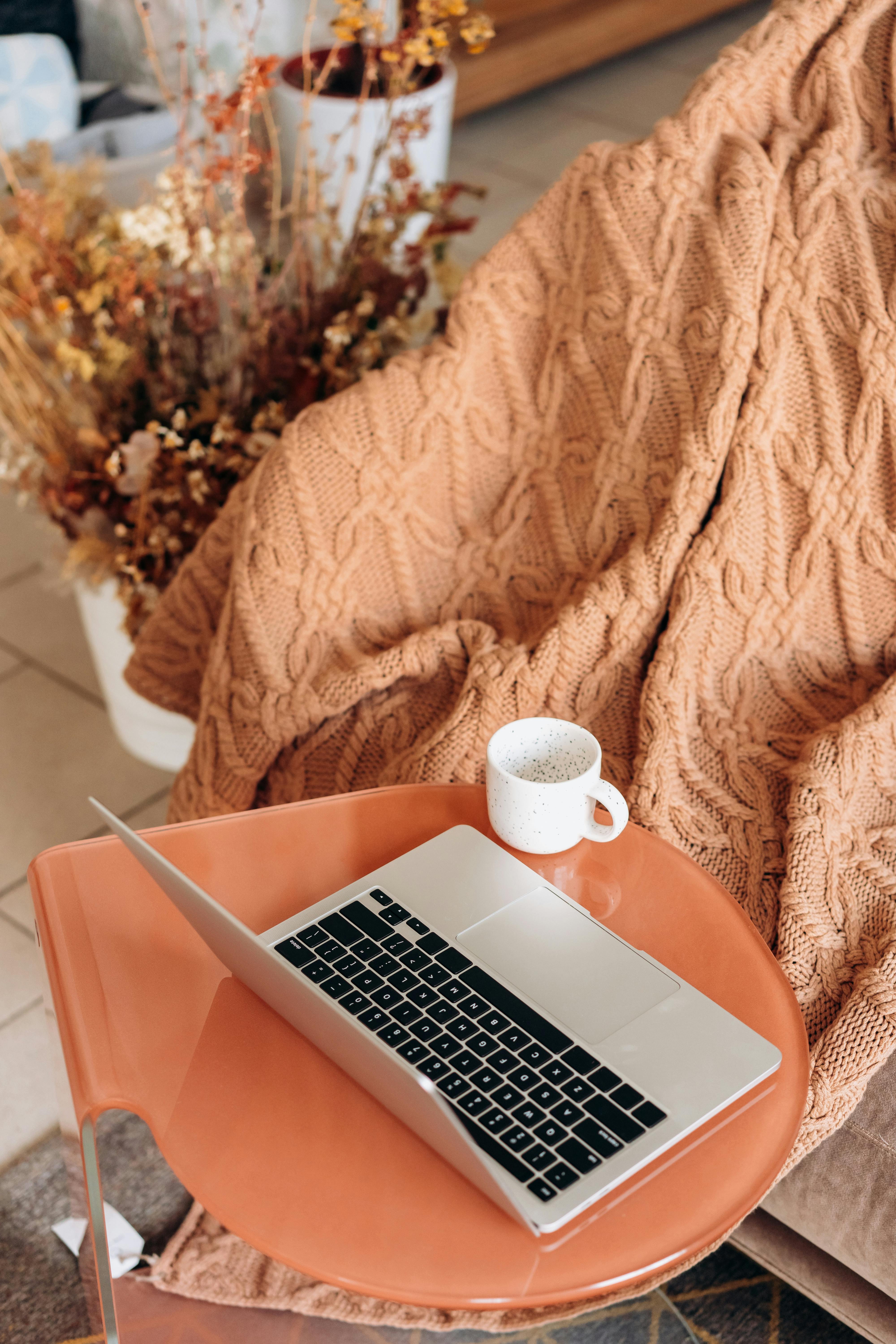 Warm indoor scene with a laptop, coffee mug, and beige knit blanket on an orange table.
