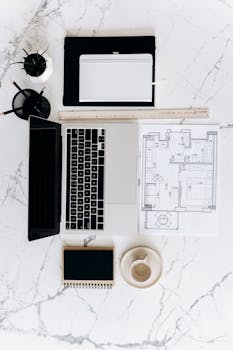 A clean, minimalist workspace setup featuring a laptop, blueprint, and coffee cup on a marble surface.