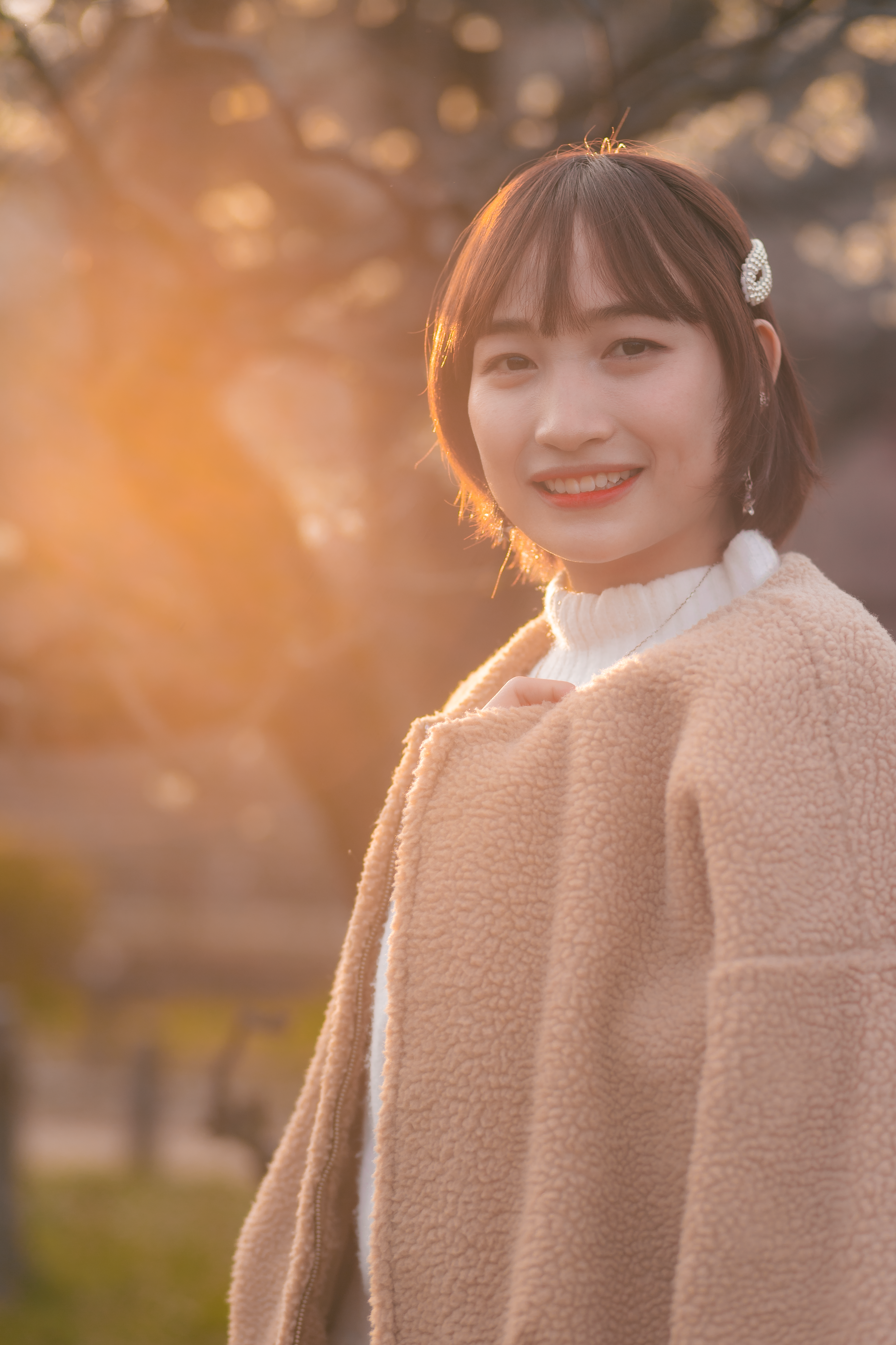 A young woman with a warm smile standing outdoors at sunset, wearing a stylish coat.