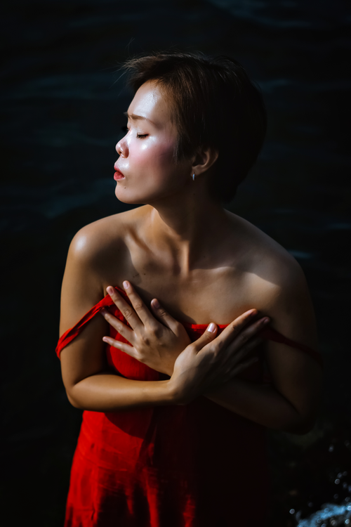 Free Serious young ethnic lady in red dress with bare shoulders and hands on chest with closed eyes near sea in sunny day Stock Photo
