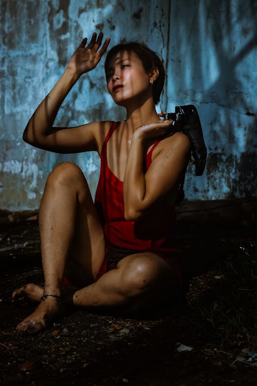 Free Full length of gentle young ethnic female in red dress sitting barefoot on soil with shoe in hand near shabby stone wall while looking away with shadows on face Stock Photo