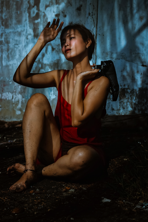Free Full length of gentle young ethnic female in red dress sitting barefoot on soil with shoe in hand near shabby stone wall while looking away with shadows on face Stock Photo