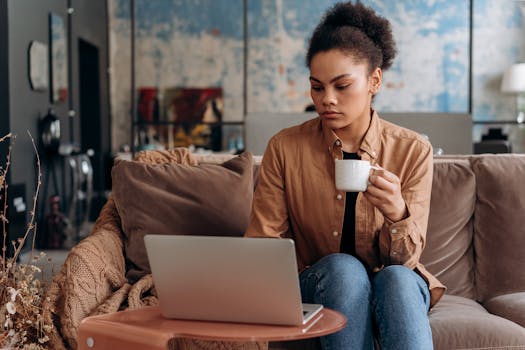 A focused black woman working from home on a laptop, holding a coffee mug, seated on a cozy couch.