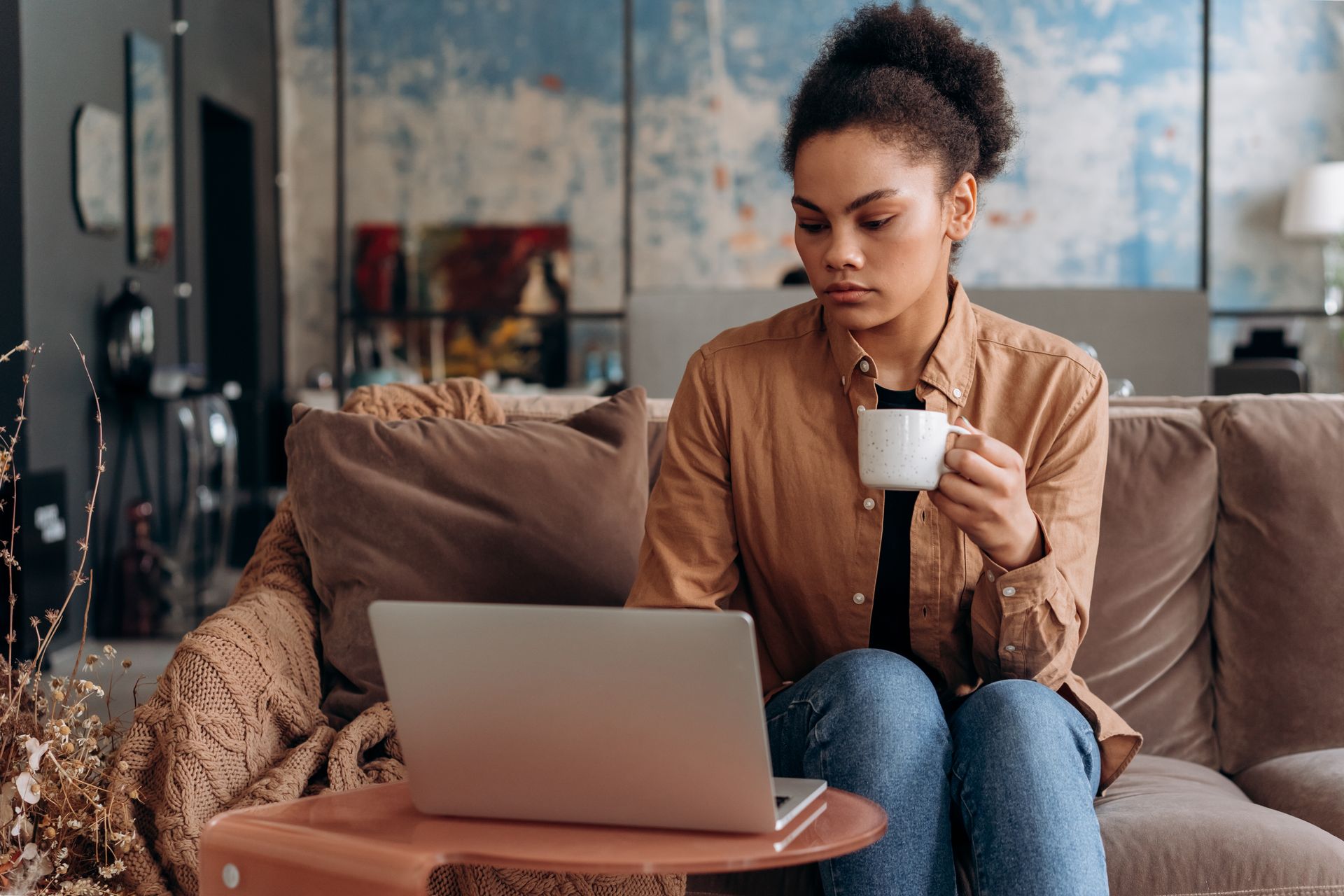 A focused black woman working from home on a laptop, holding a coffee mug, seated on a cozy couch.