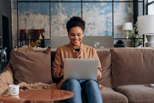 Free Smiling woman sits on a couch using a laptop, enjoying her home environment. Stock Photo