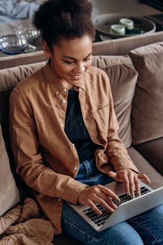 African American woman sitting on sofa, typing on a laptop, working from home.