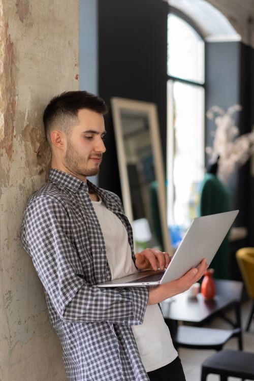 Free Stylish young man leaning on wall while working on a laptop at home. Stock Photo