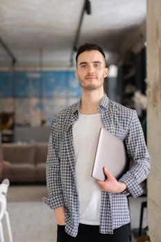 Young man in checkered shirt holding a laptop in a contemporary office setting.