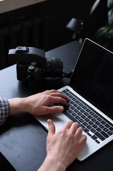 A person working on a laptop with a camera nearby on a black desk.