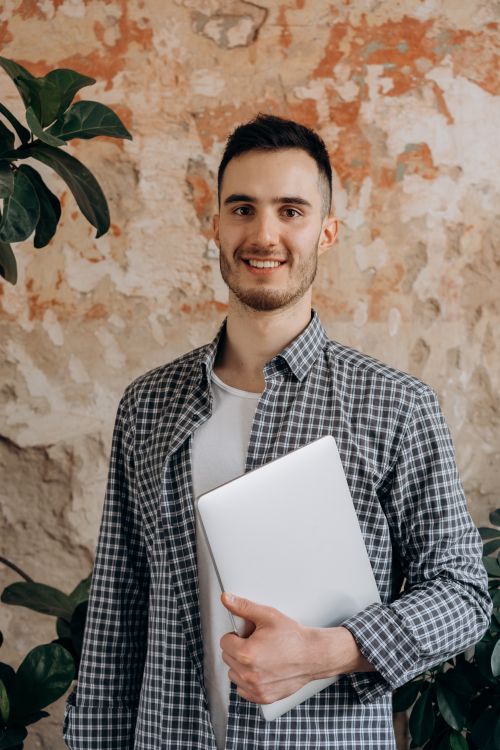Free Caucasian man smiling while holding a laptop, surrounded by a rustic indoor setting. Stock Photo