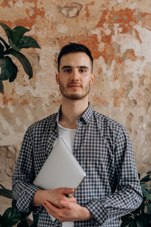 Free Young man with beard holding a laptop in front of a textured wall indoors. Stock Photo
