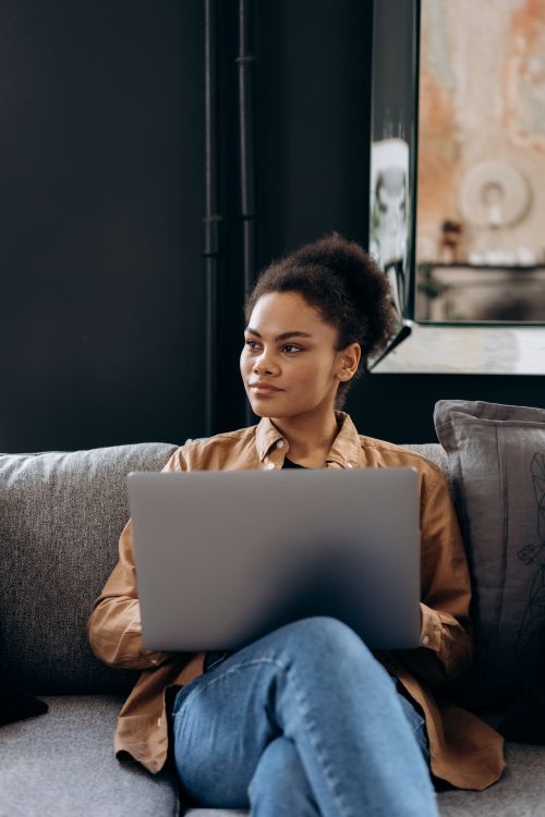 Free African American woman sitting on a sofa using a laptop, looking away thoughtfully. Stock Photo