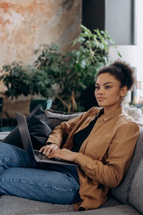 Free African American woman sitting on a sofa, working on a laptop in a cozy living room setting. Stock Photo