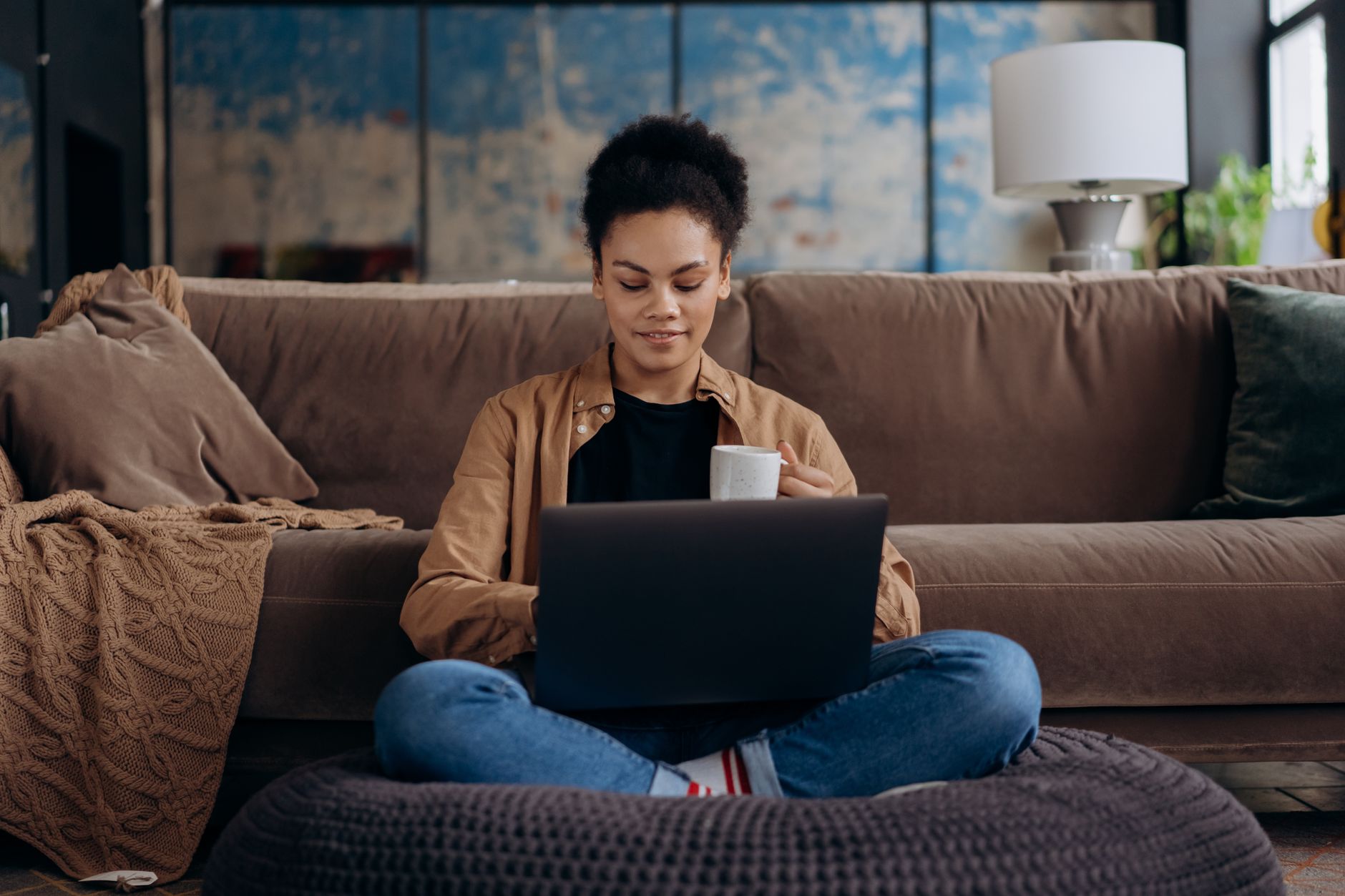 Woman sitting on a sofa with a laptop and cup, enjoying relaxed remote work indoors.