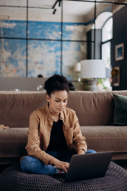 Free A woman sits on a floor cushion, working on a laptop in a modern living room. Stock Photo