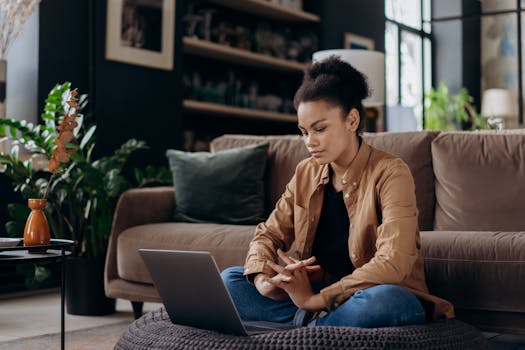 Young woman with afro hair sits on bean bag working on laptop from home, in a cozy living room setting.