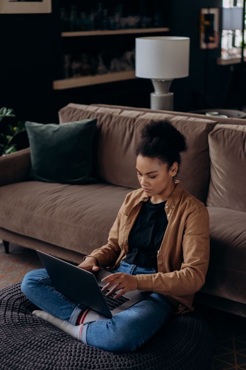Free African American woman sitting cross-legged on floor, working remotely on laptop. Stock Photo