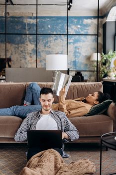 Interracial couple relaxing at home, one using a laptop, the other reading on a couch.