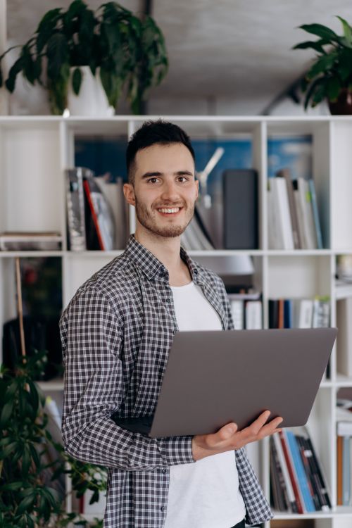 Free Smiling young caucasian man in office with laptop, indoor plants, and bookshelves. Stock Photo