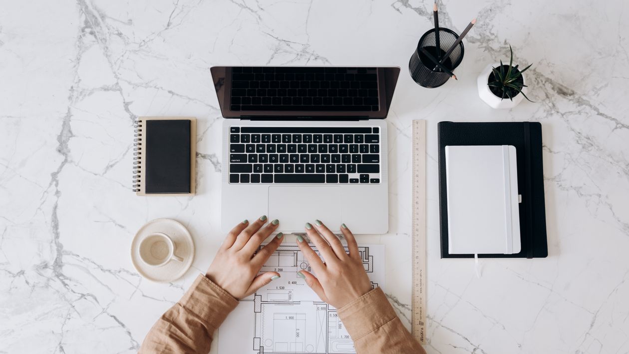 Free Top view of a stylish home office desk with a laptop, planner, and coffee cup, showing hands on a blueprint. Stock Photo