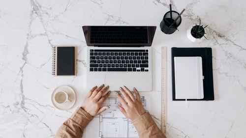 Free Top view of a stylish home office desk with a laptop, planner, and coffee cup, showing hands on a blueprint. Stock Photo