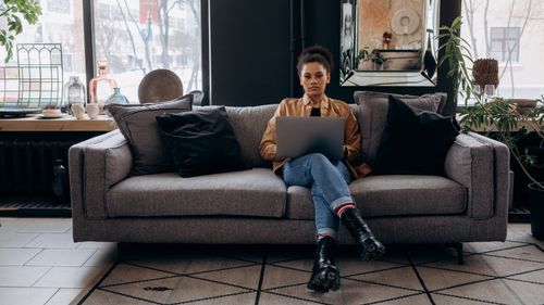 Free Young woman seated on a comfortable gray sofa using a laptop, surrounded by indoor plants. Stock Photo