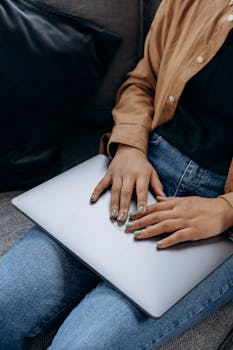 A woman sitting on a sofa with a laptop on her lap, wearing a brown jacket and denim pants.