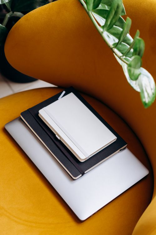 Free Stylish workspace featuring notebooks and a laptop on a mustard armchair adorned with green leaves. Stock Photo