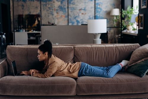 Free African American woman lying on sofa using laptop for remote work at home. Stock Photo