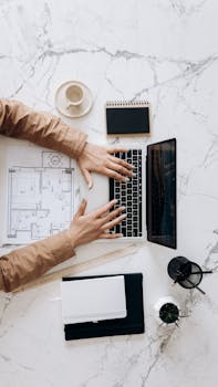 Overhead view of architect using laptop with blueprints and coffee on marble desk.