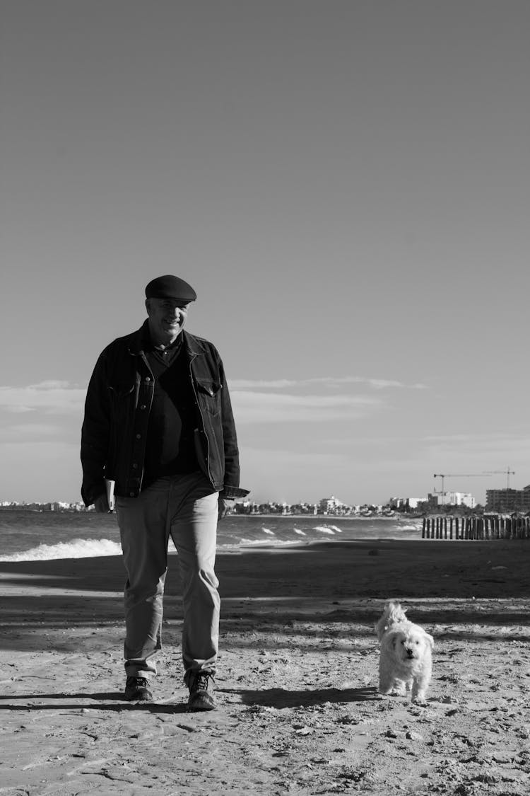 
A Grayscale Of A Man Walking On A Beach With His Pet Dog