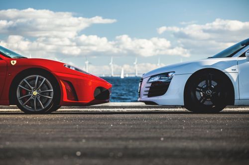 Free Red Ferrari and white Audi R8 facing each other with wind turbines in the background by the sea. Stock Photo