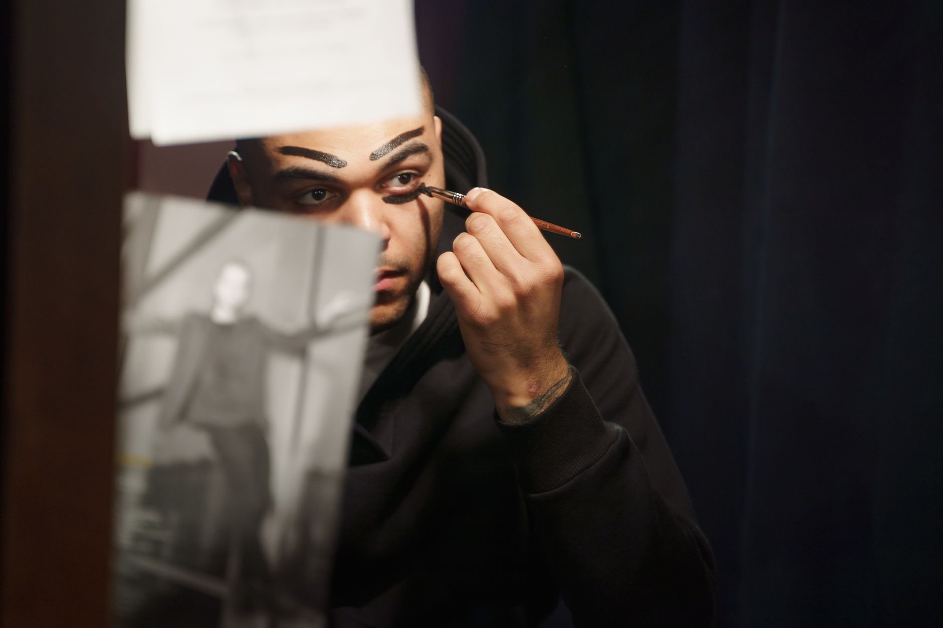 A male actor applies makeup in a theater dressing room, preparing for a performance.