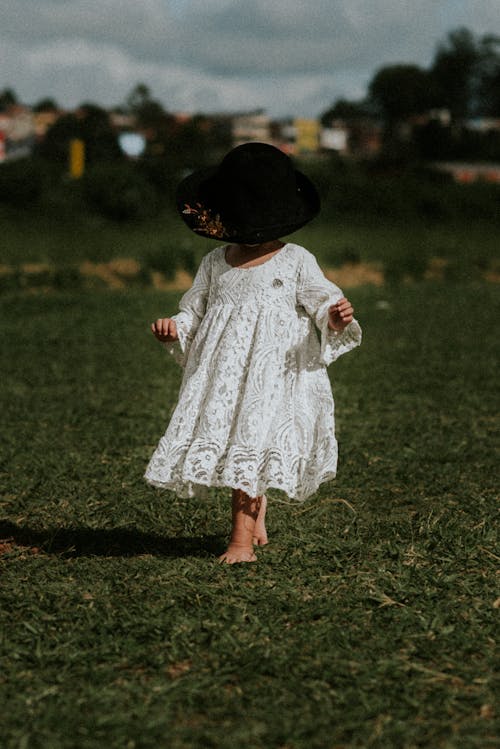 Little Girl in White Dress and Black Hat Walking on Green Grass Field