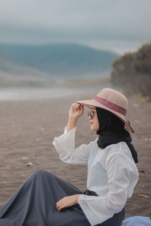 Free Woman in hijab, wearing a hat, sits on a beach enjoying the serene view. Stock Photo